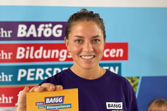 Eine junge Frau steht vor einem Plakat-Banner und zeigt die Postkarte der BAfög-Tour ins Bild. Darauf steht "Meht Bafög, mehr Bildungschancen, mehr Zukunft, mehr für dich.